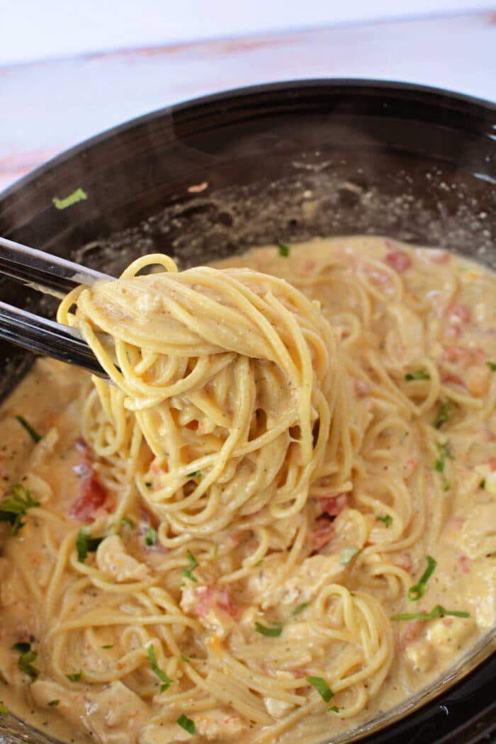 Tongs lifting creamy chicken spaghetti with tomatoes and herbs from a black bowl, fresh from the crockpot.