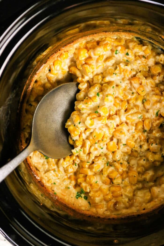 A spoon in a Crockpot filled with creamy cooked corn, garnished with herbs—solid for savoring good Crockpot Creamed Corn.