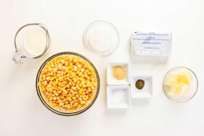 Bowls of corn, butter, seasonings, cream, and sugar on a white surface, ready for cooking the solid Crockpot Creamed Corn.