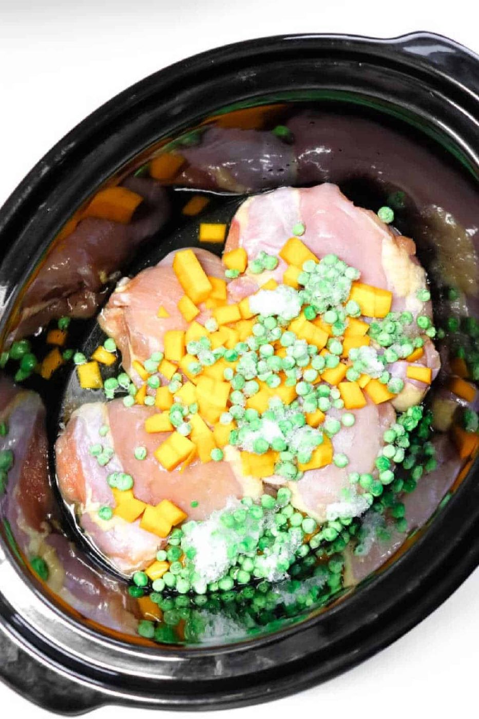 Raw chicken, frozen peas, and diced yellow vegetables in a black slow cooker, solid for making wholesome Crockpot Dog Food.