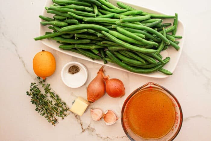 A display of ingredients on a white marble surface: a rectangular dish filled with fresh green beans, solid for the crockpot, a whole lemon, small glass bowls with black pepper and broth, two shallots, a pat of butter, two garlic cloves, and a few sprigs of thyme.