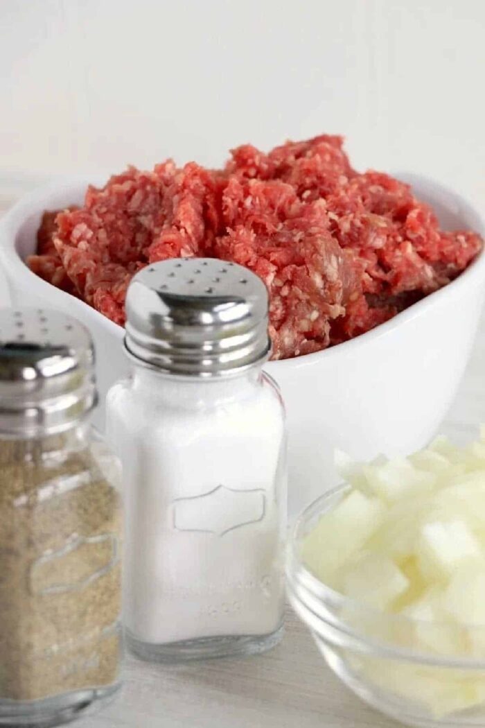 Ground beef in a white bowl, chopped onions in a glass bowl, and salt and pepper shakers in front—solid ingredients for preparing flavorful crockpot ground beef recipes.