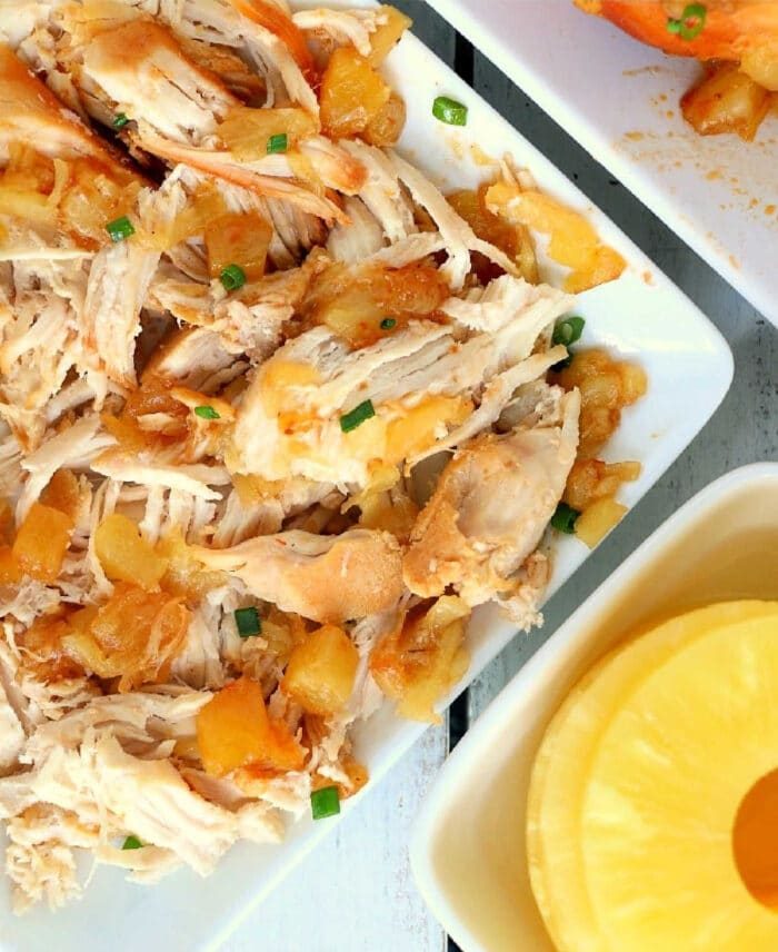 Crockpot Hawaiian chicken, featuring tender shredded pieces, is beautifully presented with pineapple chunks and green onions on a white plate.