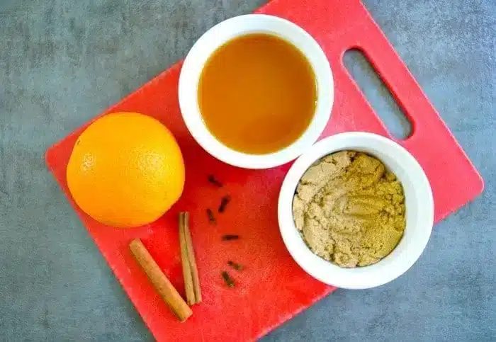A red cutting board holds an orange, a small bowl of brown sugar, a small bowl of liquid, cinnamon sticks, and cloves on a gray surface—solid ingredients for crafting crockpot hot apple cider.
