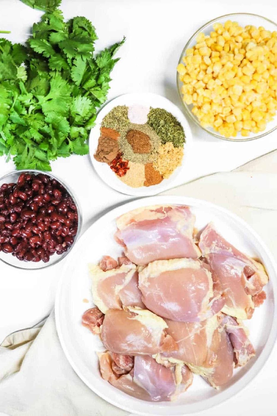 Raw chicken thighs, black beans, corn, cilantro, and assorted spices arranged in bowls on a white surface—solid for making flavorful crockpot jerk chicken.