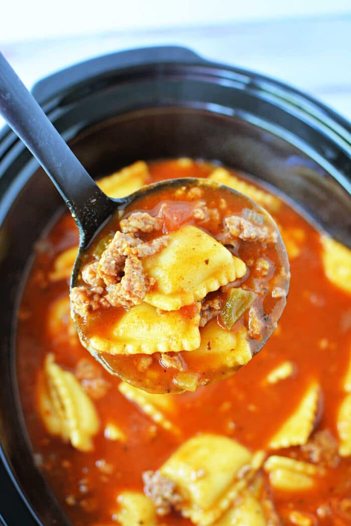 A ladle holds ravioli, ground beef, and tomato sauce over a Crockpot filled with the same mixture, creating a comforting Crockpot Lasagna Soup.