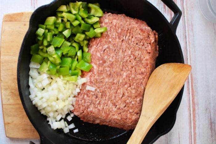 Ground meat, chopped green bell pepper, and chopped onion in a cast iron skillet with a wooden spatula—solid for starting your Slow Cooker Lasagna Soup.
