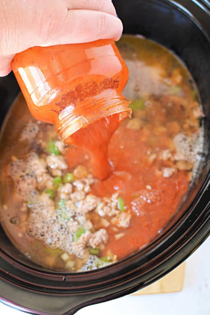 A hand pours tomato sauce from a jar into a Crockpot filled with ground meat and vegetables, solid for making hearty Lasagna Soup.