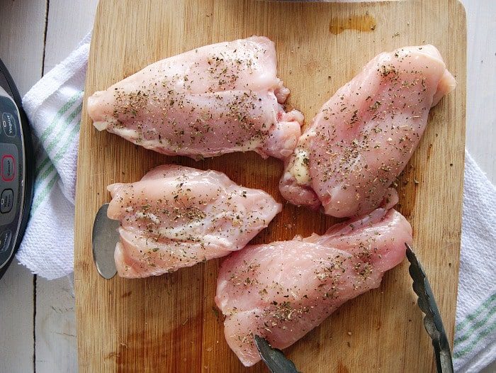 Four seasoned raw chicken thighs, solid for a crockpot lemon pepper chicken recipe, rest on a wooden cutting board alongside a trusty pair of tongs.