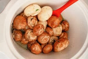 Crockpot mushrooms seasoned with herbs rest in a white slow cooker, with a red spatula nestled inside.