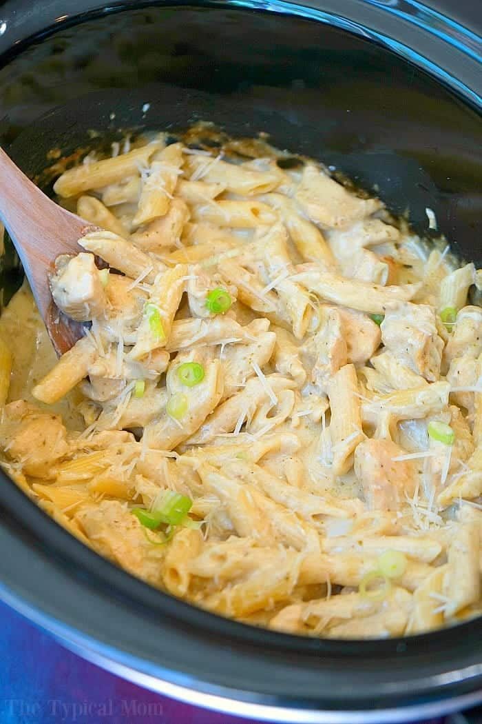 Creamy crockpot pesto chicken alfredo casserole with penne, cheese, and green onions, all blended together in a slow cooker with a wooden spoon.