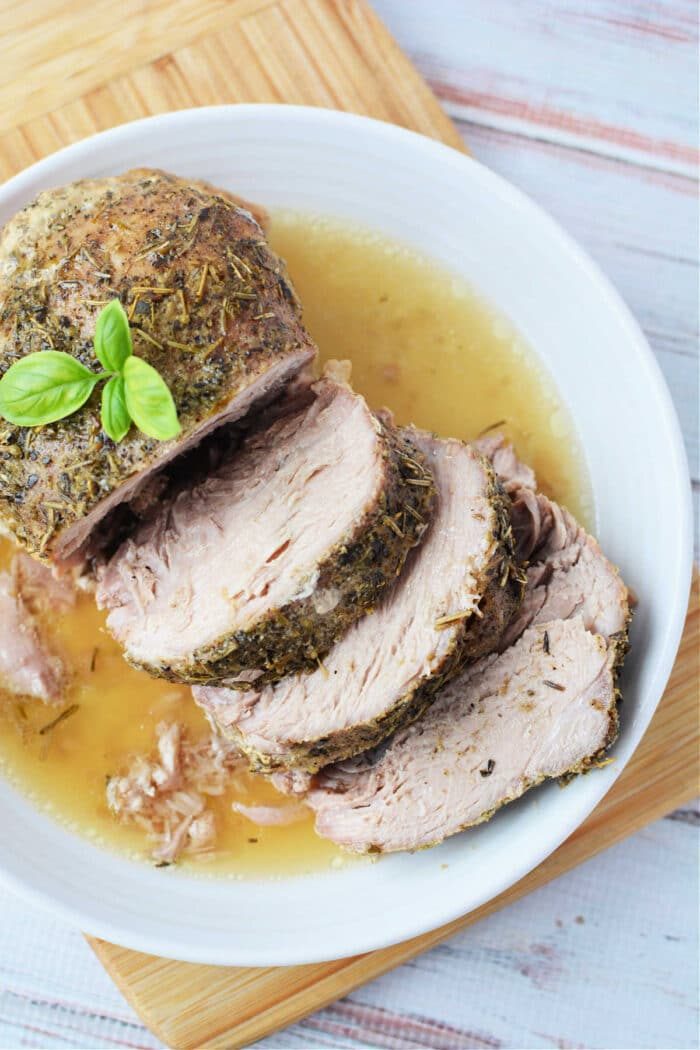 Enjoy a deliciously tender Crockpot Porketta Roast, perfectly herb-crusted and sliced, served with rich gravy on a white plate. Garnished with fresh basil for an added touch of flavor.