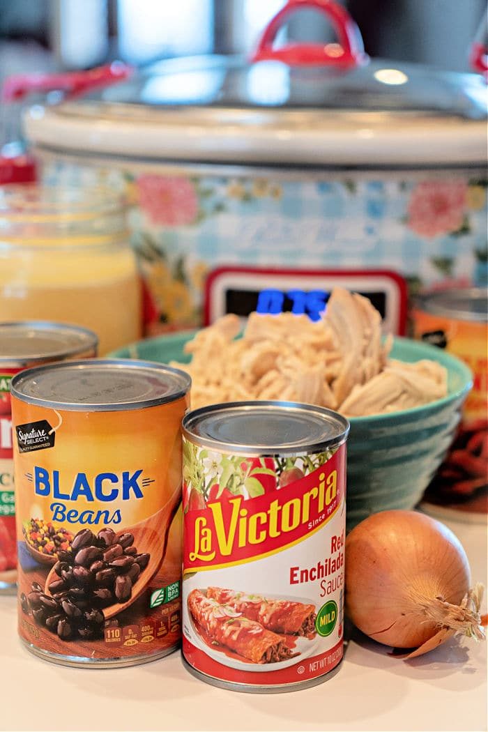 Canned black beans, enchilada sauce, an onion, and a slow cooker blend seamlessly to create an easy enchilada soup. The dish is ready to be served with shredded chicken in a bowl nearby.