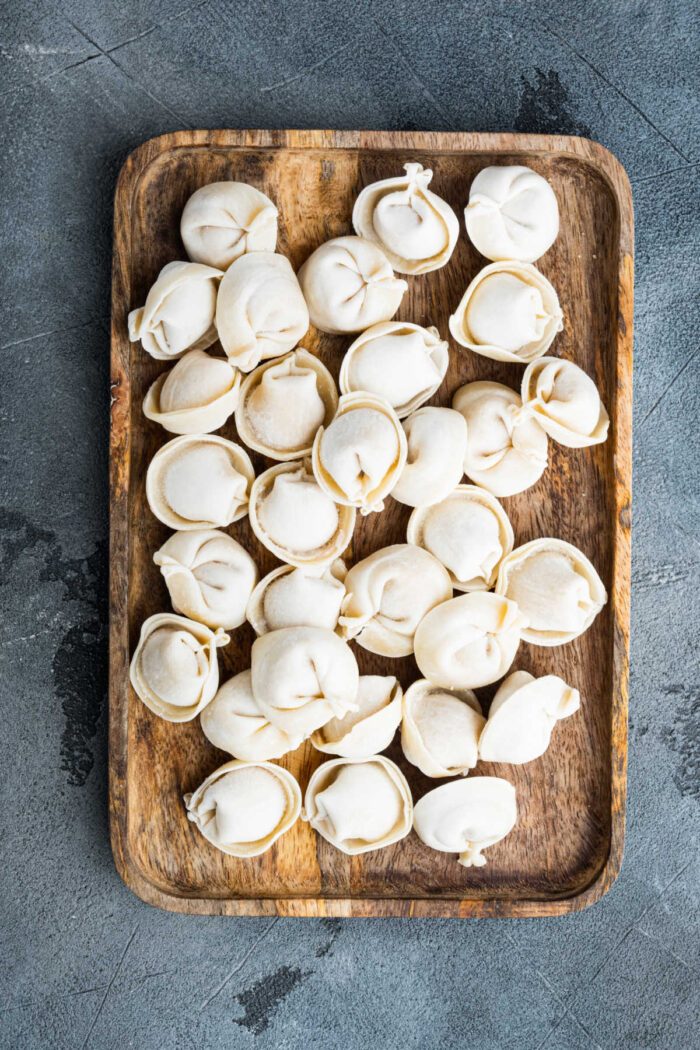 A wooden tray filled with uncooked dumplings on a textured gray surface, solid inspiration for those exploring Crockpot Recipes with Tortellini.