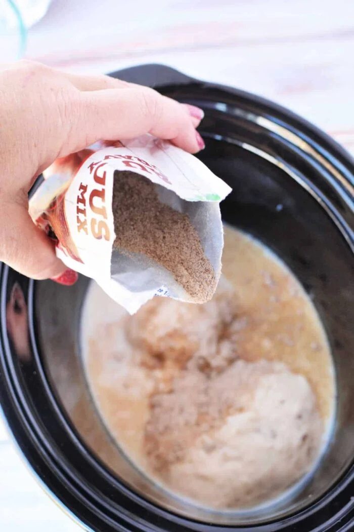 A hand pours dry biscuit mix from a packet into a slow cooker with liquid ingredients inside, preparing for a good Crockpot Salisbury Steak meal.