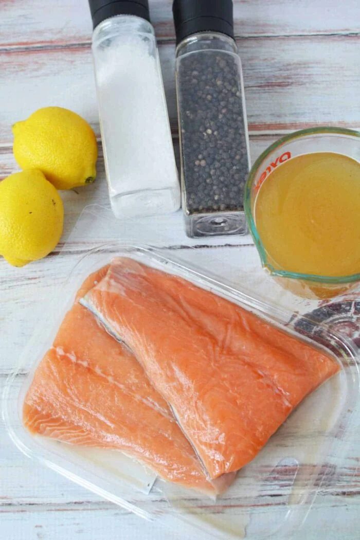 Raw salmon fillets in a plastic tray with lemons, salt, pepper, and a measuring cup of broth—solid ingredients for easy Crockpot Salmon on your table.