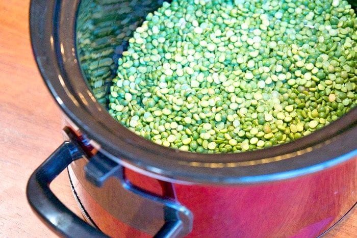 A slow cooker brimming with uncooked green split peas and savory ham rests on a wooden surface, ready to become a hearty split pea soup.