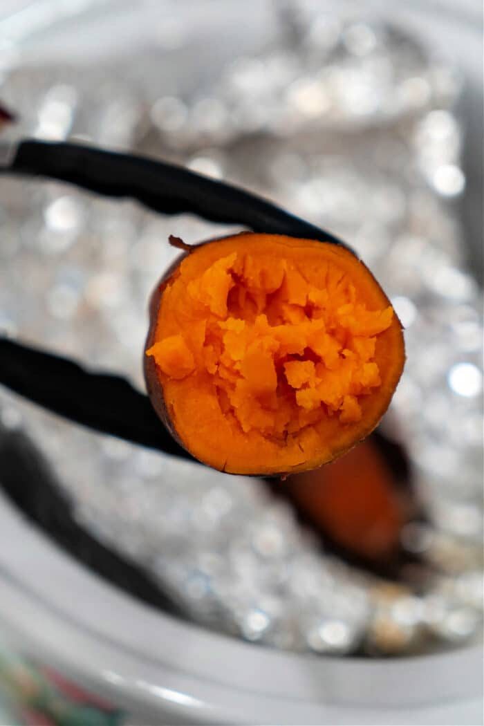 A cooked sweet potato held by tongs, with vibrant orange flesh visible against a blurred kitchen backdrop, showcases the delightful simplicity of crockpot sweet potatoes.