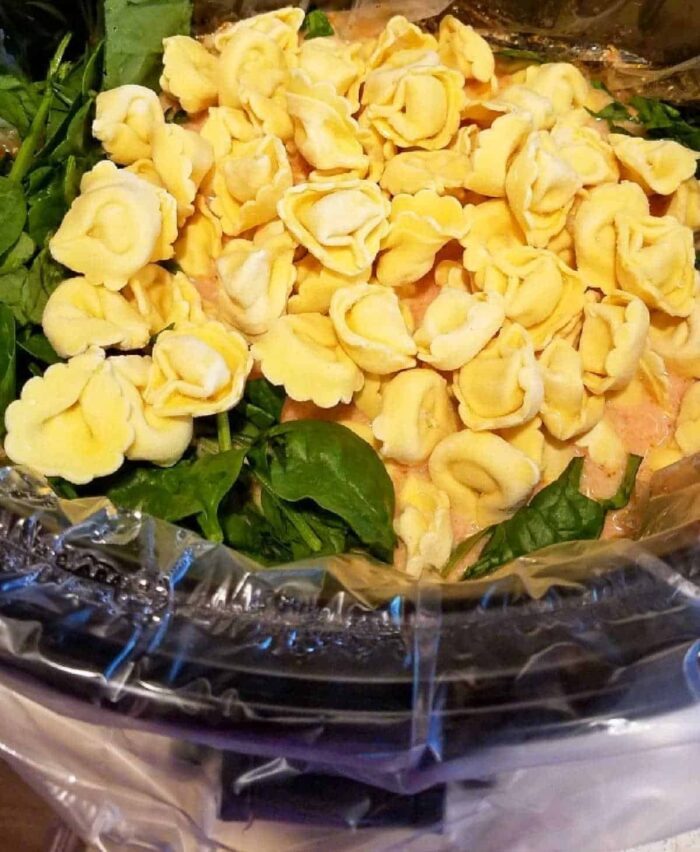 Uncooked tortellini pasta and fresh spinach in a slow cooker lined with a plastic liner—solid for starting your crockpot tortellini soup.