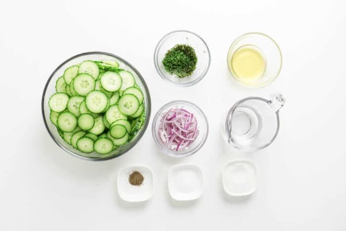 Sliced cucumber, red onion, fresh dill, vinegar, water, salt, pepper, and sugar in bowls on a white background—solid ingredients for a refreshing cucumber salad.
