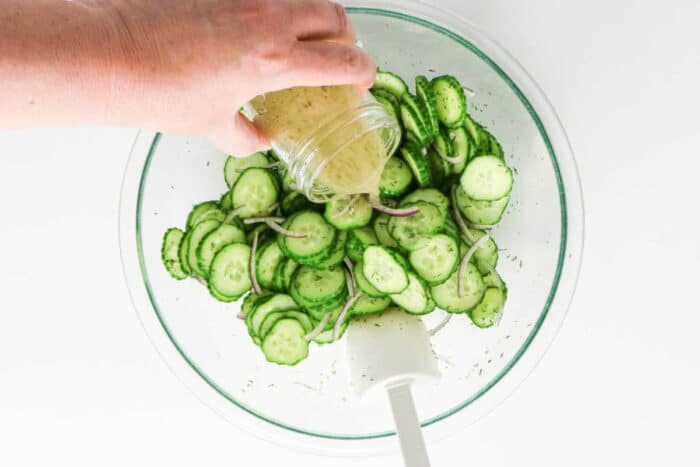 A hand pours dressing over sliced cucumber and onion in a glass bowl with a white spatula, creating a fresh salad solid for adding dill.