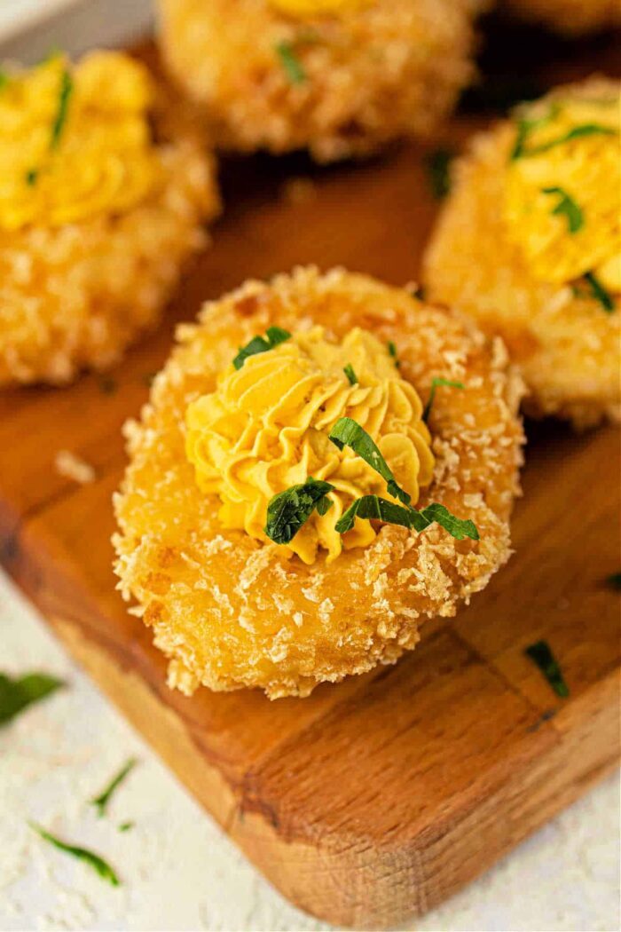 A crispy breaded deep-fried deviled eggs, topped with a yellow piped sauce and garnished with green herbs on a wooden board.