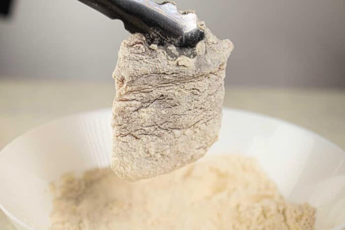 A piece of breaded boneless pork chops, soon to become a crispy delight, is held by tongs over a bowl of flour, ready to transform into savory deep-fried pork chops.