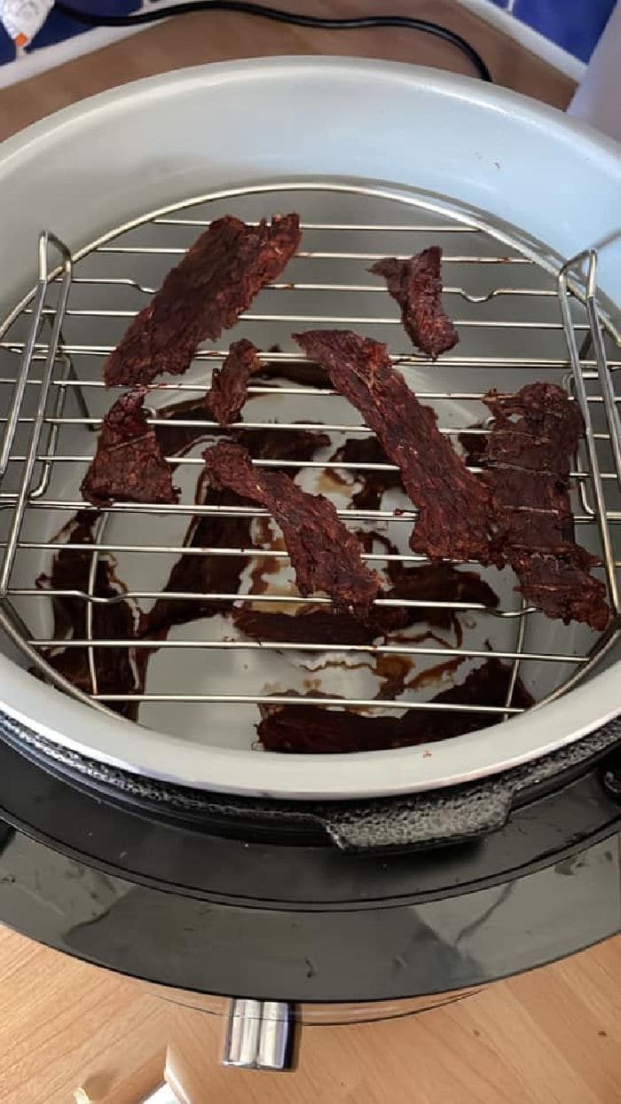 A round dehydrator with a metal rack inside holds several evenly spaced strips of dark brown dehydrated dog treats. The interior base of the dehydrator is marked with drips of liquid residue.