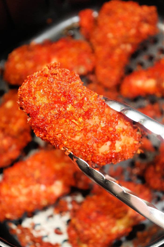 Tongs hold a piece of breaded and fried chicken, seasoned to perfection with Doritos Ranch Dipped Hot Wings spices, surrounded by more tantalizing pieces in the background.