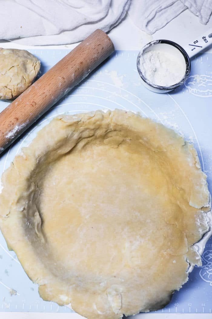 Unbaked pie crust in a pan, with a rolling pin, dough ball, and cup of flour nearby on a blue surface—solid foundations for a homemade Double Crust Pot Pie.