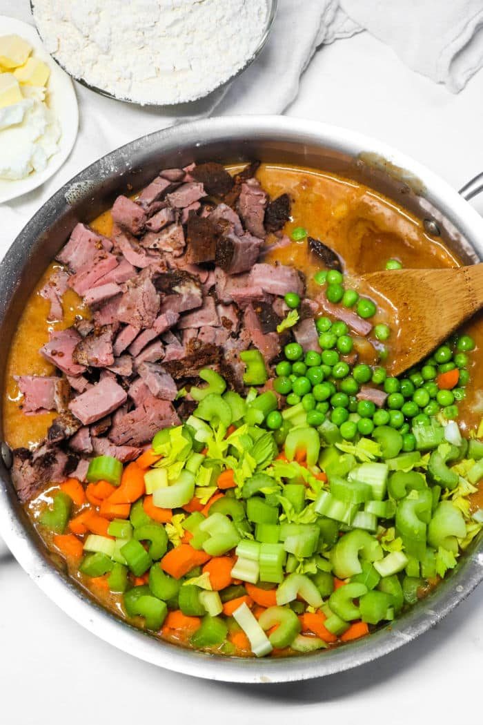 A skillet with diced meat, peas, chopped celery, carrots, and creamy sauce being stirred with a wooden spoon—the solid filling for a comforting Double Crust Pot Pie.