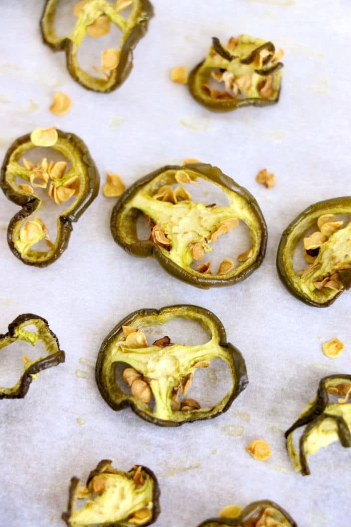 Sliced pieces of dried jalapeño peppers are scattered on a sheet of parchment paper. The pepper slices are roasted and browned, with seeds visible in the center of some slices. Small bits of roasted pepper seeds are also present on the paper.