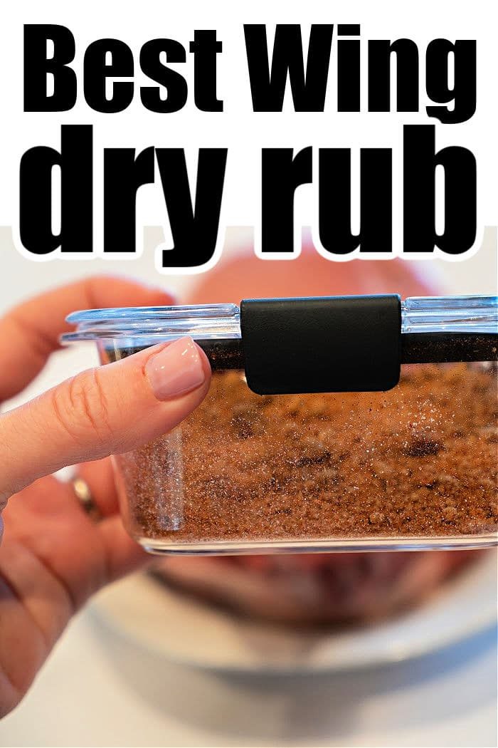 A hand holding a glass container with a dark dry rub mixture perfectly crafted for chicken wings. The container, secured with a black latch on its lid, boasts the text: "Best Wing dry rub." In the blurred background, a plate subtly awaits the masterpiece.