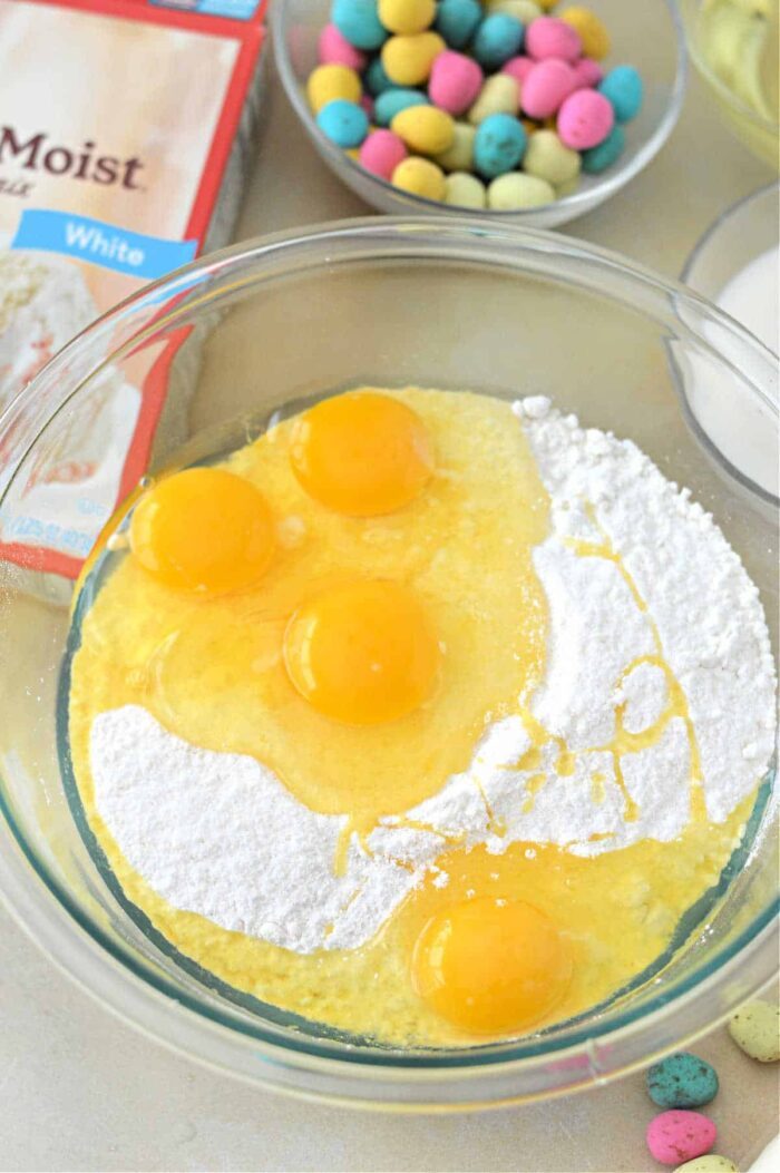 Four eggs and cake mix in a glass bowl, with an Easter Bundt Cake mix box and candy eggs in the background.