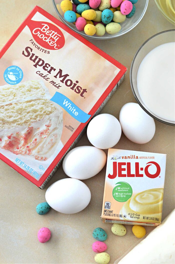 Box of cake mix, eggs, vanilla pudding mix, milk, and colorful candy eggs on a light surface—everything you need for an Easter Bundt Cake.