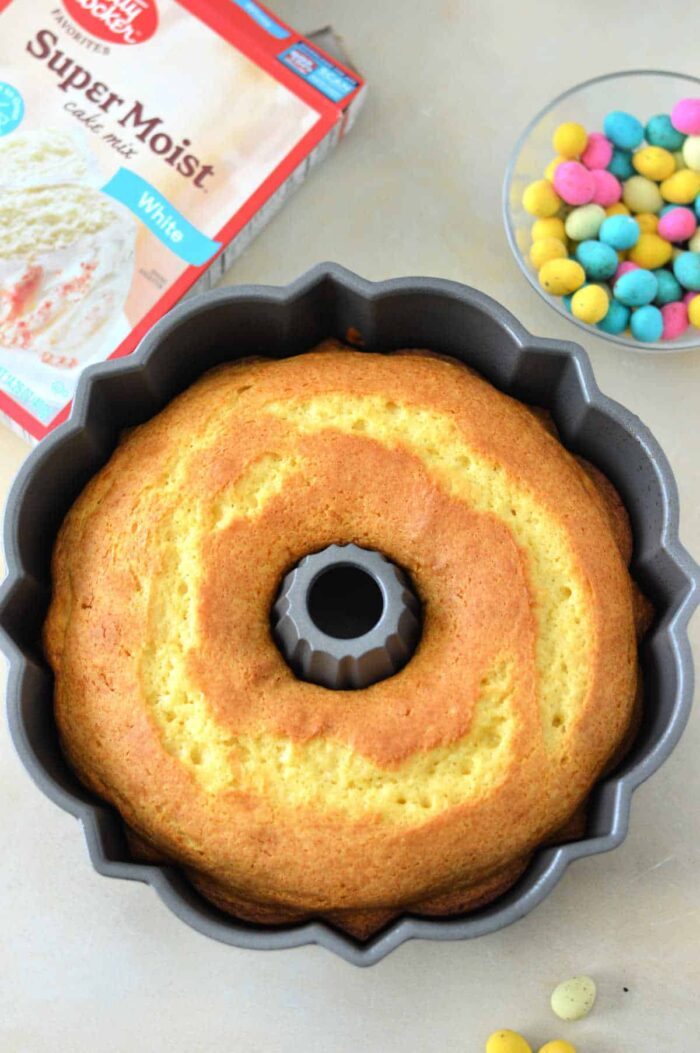 An Easter Bundt Cake sits baked in a pan, with a cake mix box and a bowl of colorful candy-coated chocolates nearby.