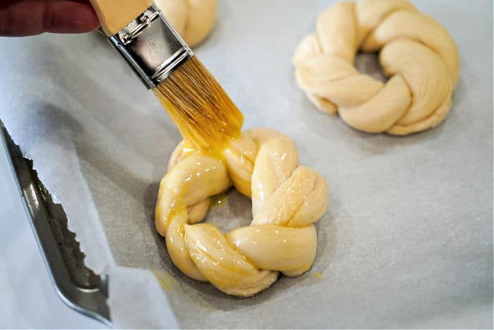 A pastry brush applies egg wash to an Easter egg bread braided dough ring on a parchment-lined baking sheet.