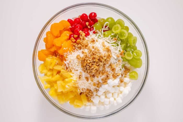 Glass bowl with grapes, pineapple, mandarin oranges, cherries, marshmallows, shredded coconut, and chopped nuts—an easy ambrosia recipe solid for any occasion.