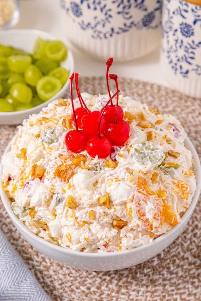 A bowl of fruit salad topped with five maraschino cherries, with grapes in the background—like an easy ambrosia recipe served fresh.