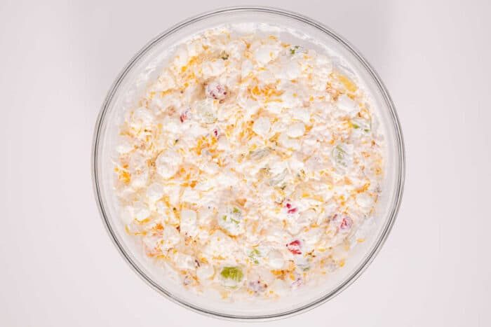 A glass bowl filled with a creamy salad mixture containing marshmallows, fruit, and shredded cheese—an easy ambrosia recipe solid for any occasion.