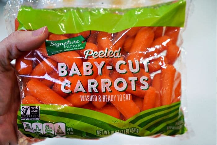 A hand holding a bag of peeled baby-cut carrots labeled "Signature Farms Quality Guaranteed." The bag states "Washed & Ready to Eat" and includes a Non-GMO Project Verified mark, solid for those wondering how to cook carrots quickly. The bag size is 16 oz (1 lb) / 454 g.