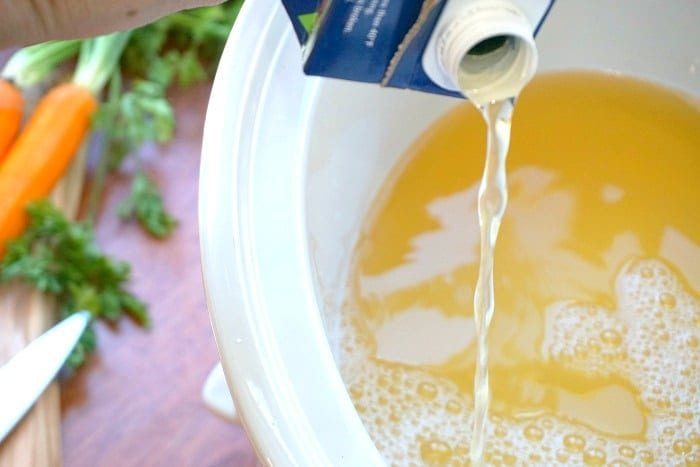 Pouring broth from a carton into a slow cooker, with carrots and parsley in the background, sets the stage for an easy Italian wedding soup recipe.
