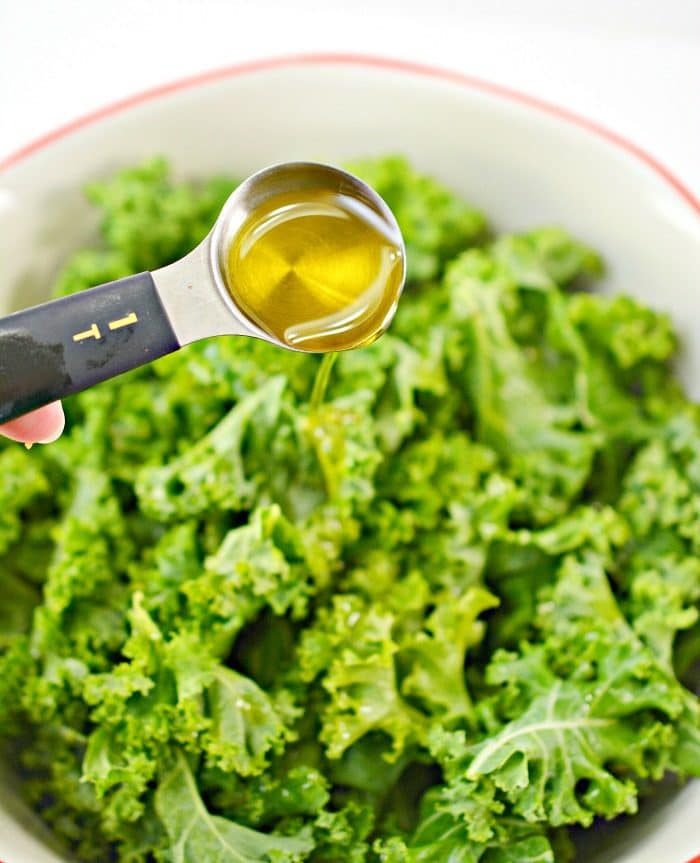 A tablespoon of olive oil is being drizzled over fresh kale in a white bowl, setting the stage for good baked kale chips.