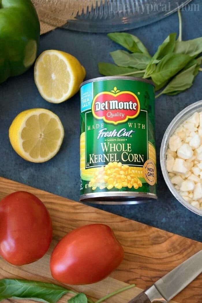 A can of Del Monte whole kernel corn surrounded by tomatoes, lemon, basil, and chopped onions—solid ingredients for a fresh Jiffy cornbread salad.