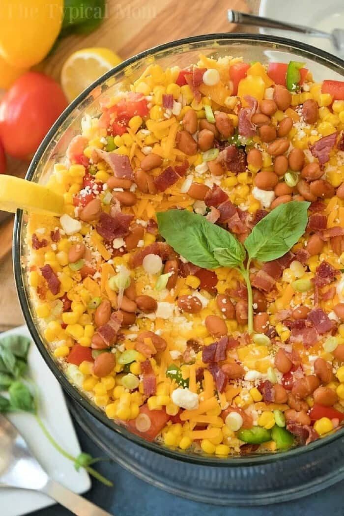Jiffy cornbread salad layered with corn, beans, cheese, bacon, green onions, and fresh basil leaves in a clear glass bowl.
