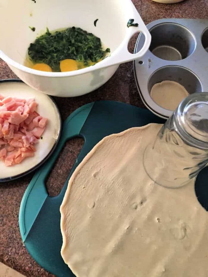 Dough, eggs with spinach, chopped bacon, and a muffin tin are prepared on a kitchen counter for this easy mini quiche recipe.