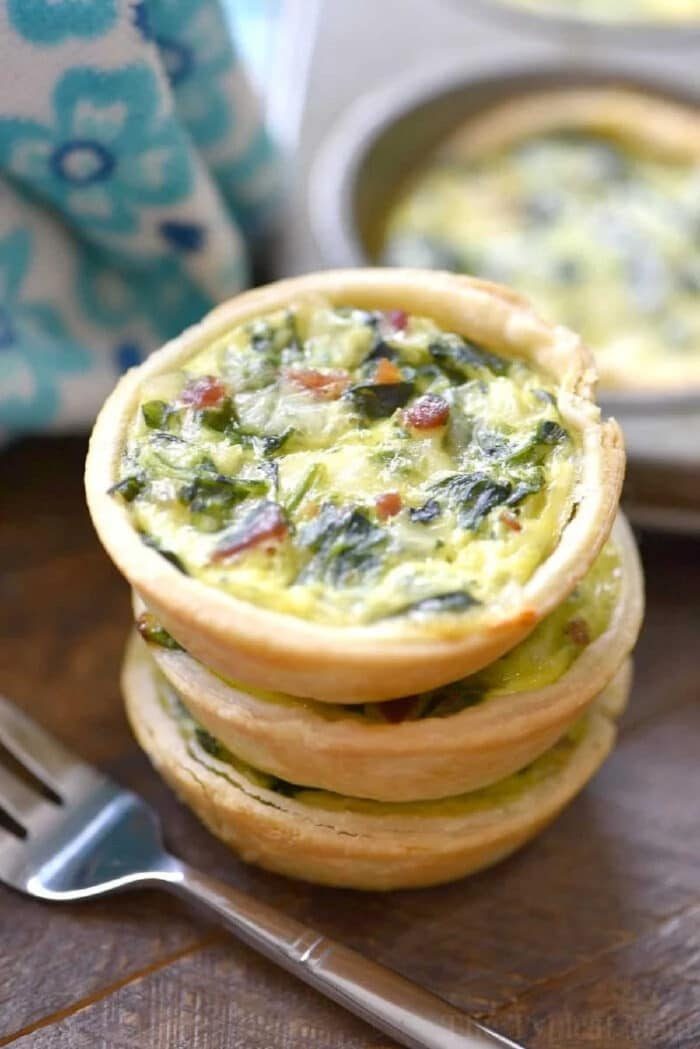 Three mini quiches made with an easy mini quiche recipe, featuring spinach and bacon, are stacked on a wooden table next to a fork and a blue patterned cloth.