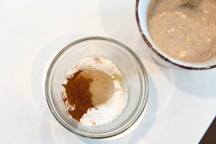 Glass bowl with flour, sugar, and cinnamon sits beside a mug containing a mixed, beige batter with a spoon—solid for an easy mug cake recipe.
