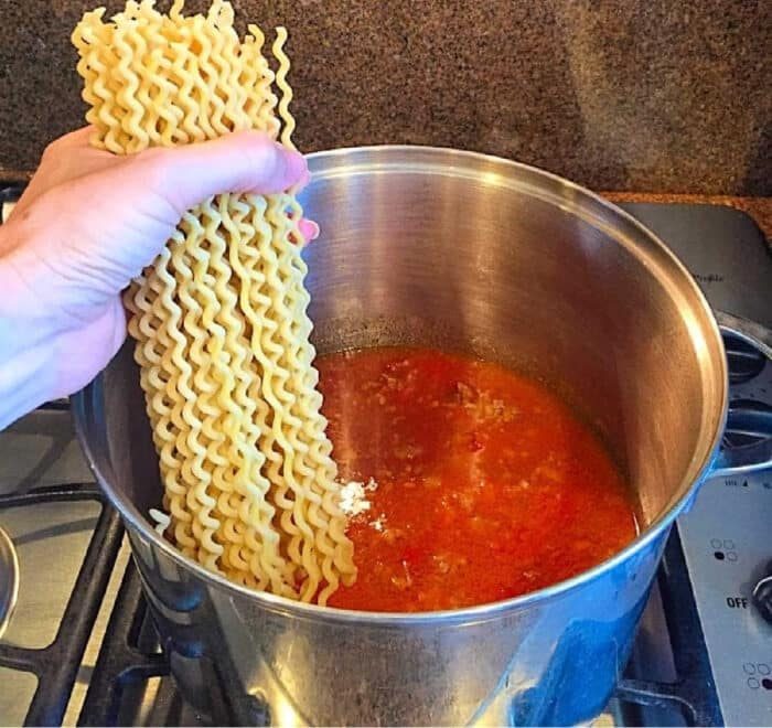 A hand holding dry curly pasta over a pot of red sauce on a stovetop, about to add the pasta for an easy one pot spaghetti meal.