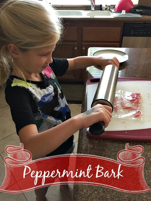 A young girl is using a rolling pin to crush peppermint candy pieces on a cutting board in a kitchen. The text "Microwave Peppermint Bark" is shown at the bottom of the image with a decorative ribbon banner.